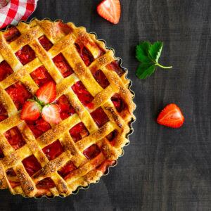 Ein ganzer Rhabarber-Erdbeer-Kuchen von oben in einer geriffelten Tarteform, mit goldbraunem Teiggitter aus quer aufgelegten Streifen, grobem Rohrzucker bestreut und roter Frucht, in der Mitte mit frischen Erdbeerscheiben garniert