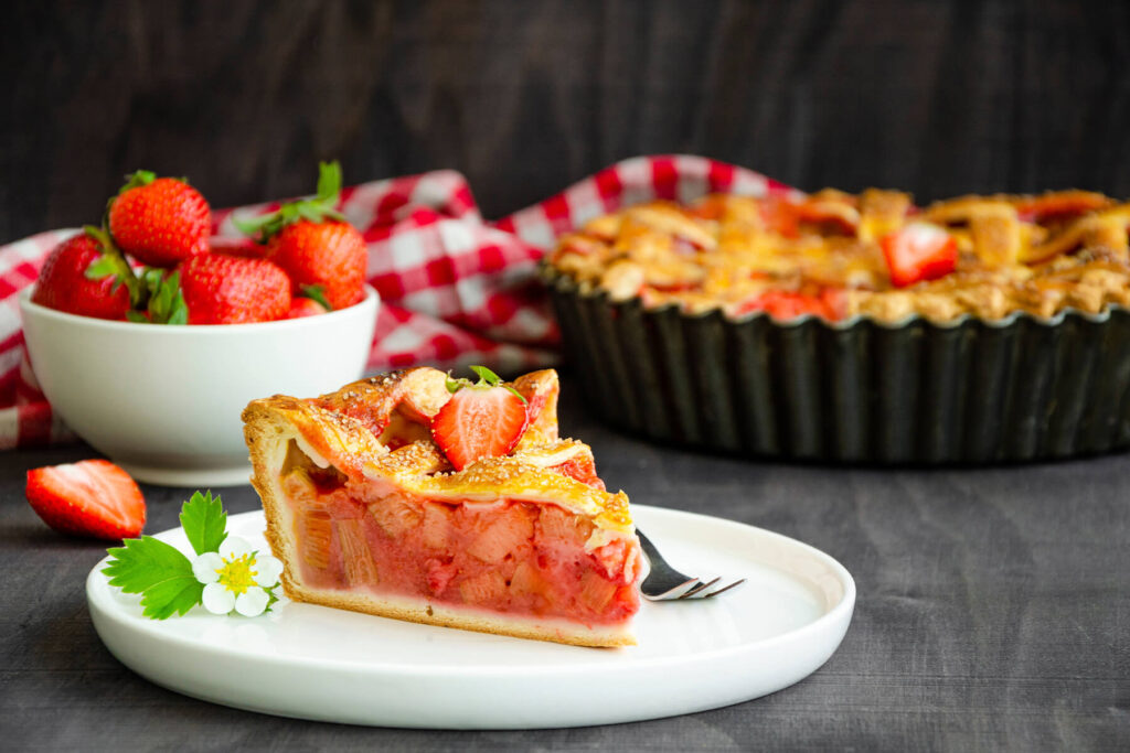 Ein Stück Rhabarber-Erdbeer-Kuchen auf einem weißen Teller, mit hellem, knusprigem Boden, rosa-roter Rhabarber-Erdbeer-Füllung mit hellen Rhabarberstücken und goldbraunem, blättrig aufgegangenem Teiggitter, garniert mit einer frischen Erdbeere, dahinter der ganze Kuchen in der Form