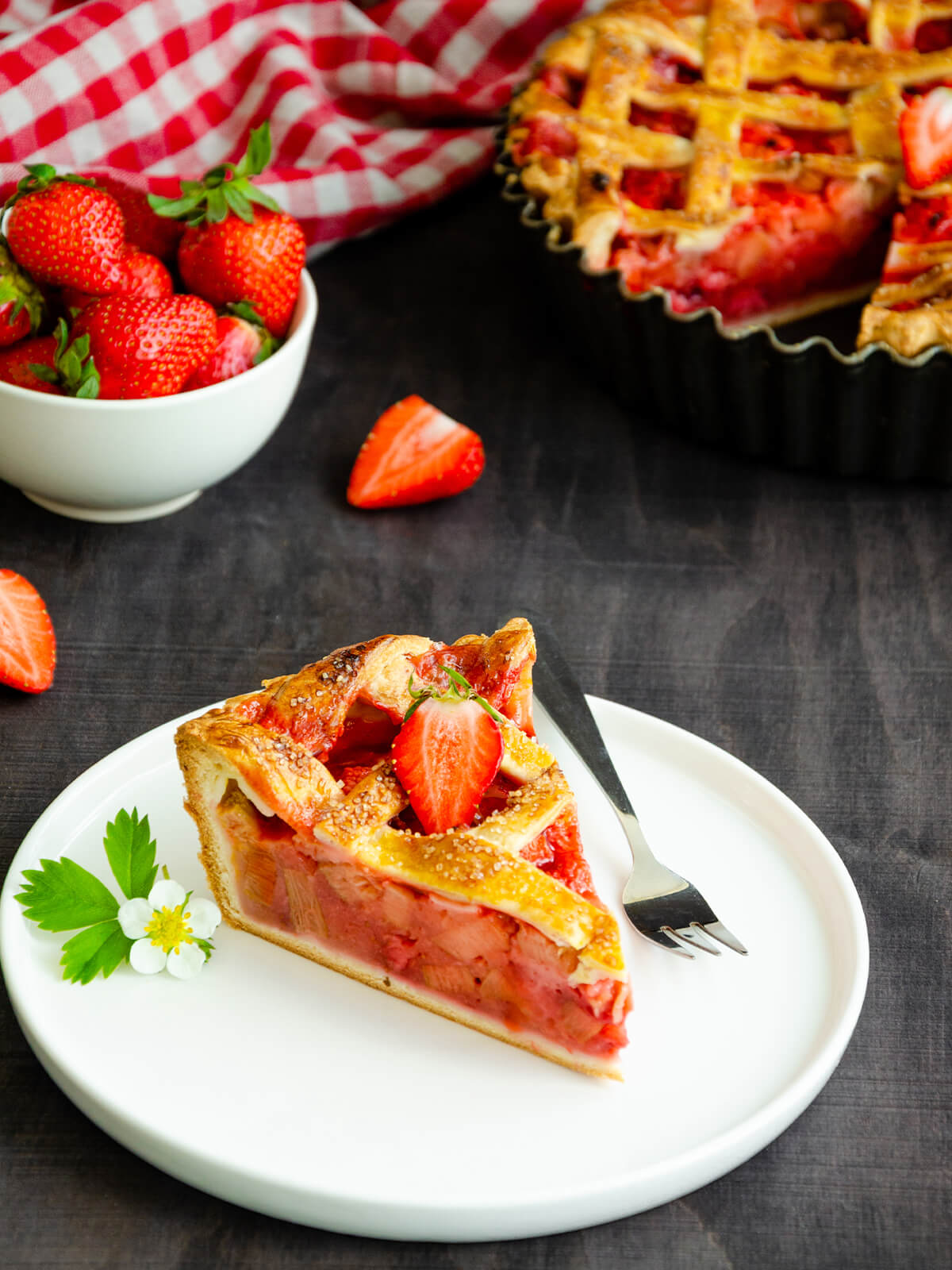 Ein Stück Rhabarber-Erdbeer-Kuchen hochkant auf einem weißen Teller, mit hellem, knusprigem Boden, rosa-roter Rhabarber-Erdbeer-Füllung mit hellen Rhabarberstücken und goldbraunem, blättrig aufgegangenem Teiggitter, garniert mit einer frischen Erdbeere und einer Erdbeerblüte, dahinter der ganze Kuchen in der Form