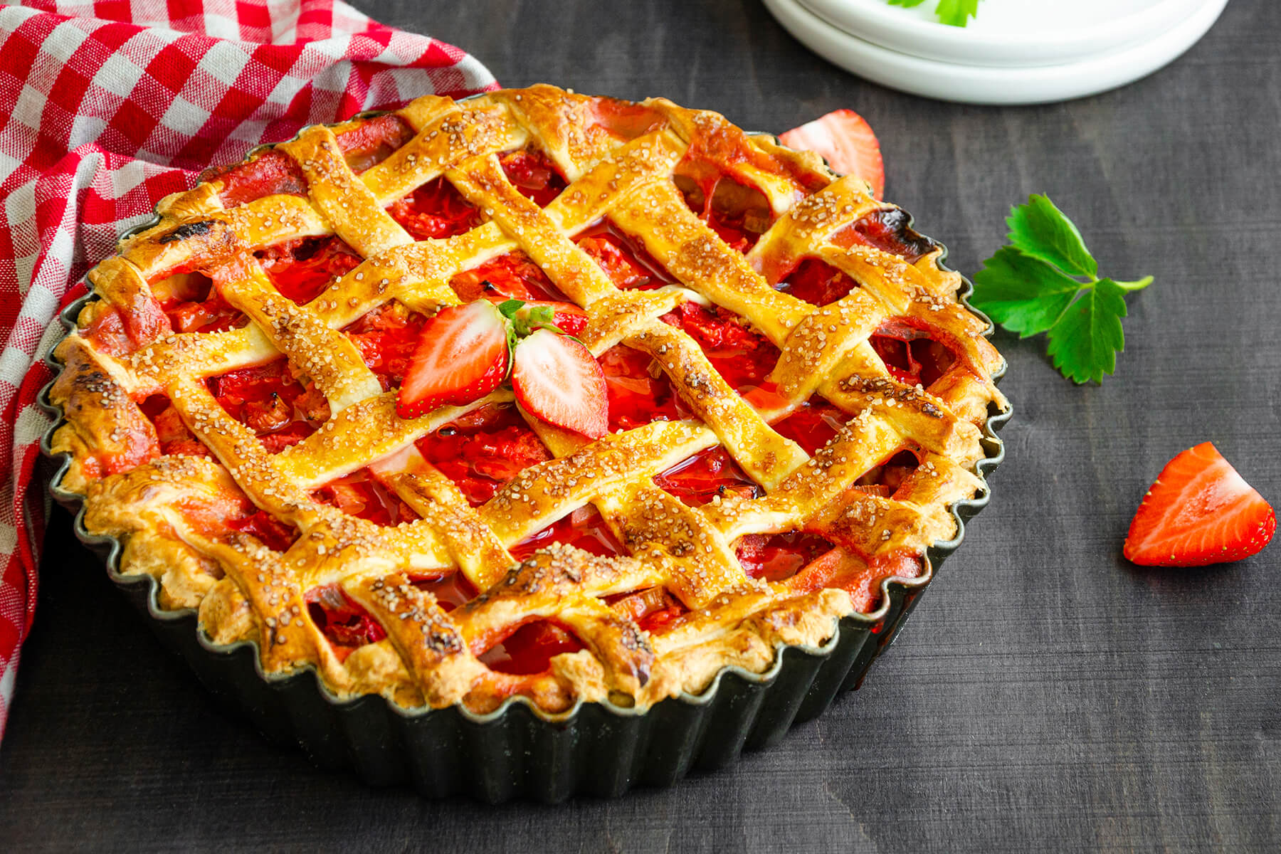 Ein ganzer Rhabarber-Erdbeer-Kuchen in einer dunklen geriffelten Tarteform, schräg von oben, mit goldbraunem Teiggitter aus quer aufgelegten Streifen, grobem Rohrzucker und roter Rhabarber-Erdbeer-Füllung, daneben ein rot-weiß kariertes Tuch