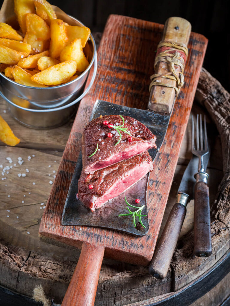 "Aufgeschnittenes Steak medium rare mit rosa Kern auf einem Hackbeil, daneben ein Becher knuspriger Steakhouse-Pommes"