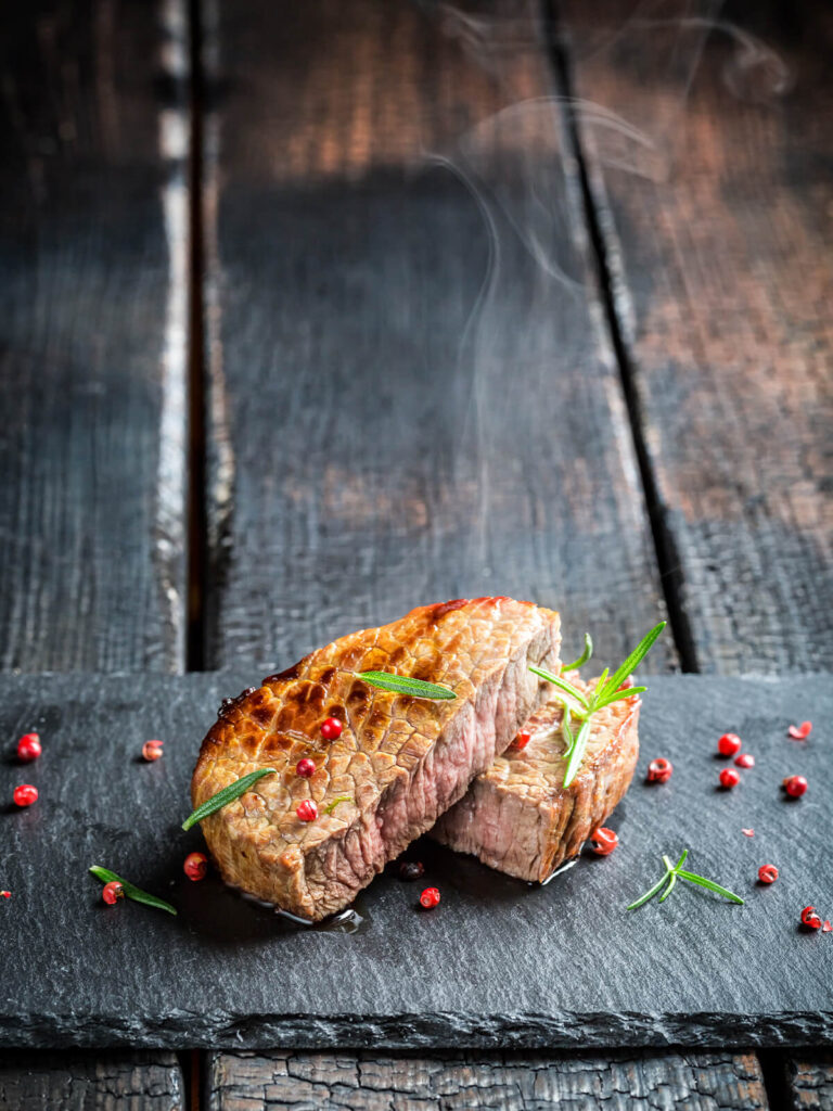 Gebratenes Rosmarin-Steak medium, aufgeschnitten mit rosa-grauem Kern, dazu Rosmarin und rosa Pfeffer auf Schiefer
