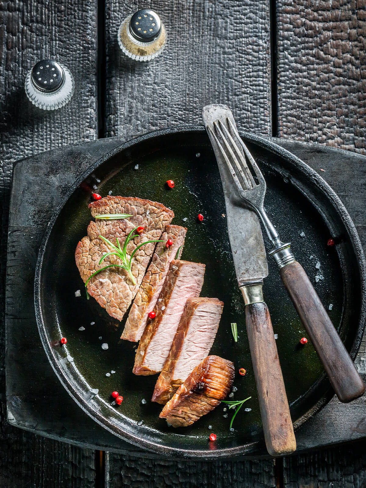 "Aufgeschnittenes Rosmarin-Steak in Scheiben auf dunklem Teller, bestreut mit rosa Pfeffer und grobem Salz"