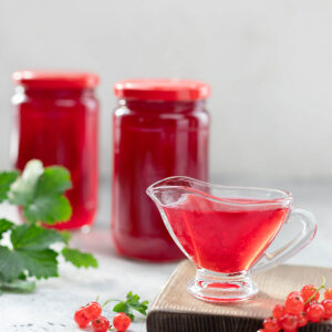 Klares rotes Johannisbeergelee in einer Glas-Sauciere, dahinter zwei gefüllte Gläser und frische Johannisbeeren