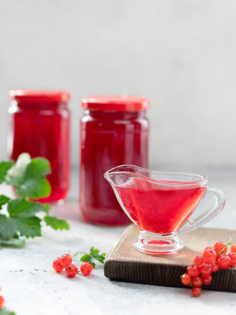Klares rotes Johannisbeergelee in einer Glas-Sauciere, dahinter zwei gefüllte Gläser und frische Johannisbeeren