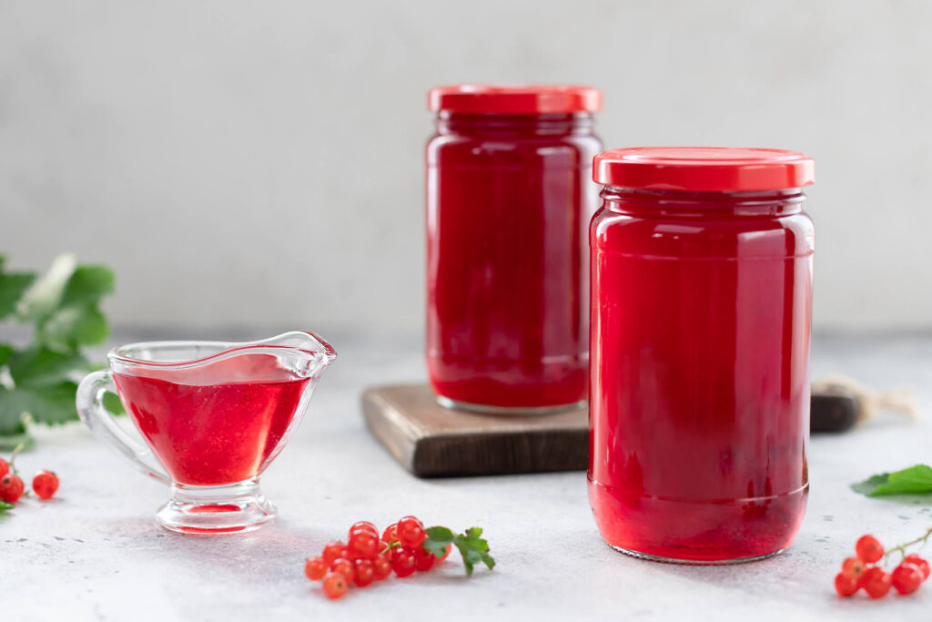 Zwei Gläser mit fertigem klarem roten Johannisbeergelee und eine Sauciere mit Gelee, daneben frische Johannisbeeren