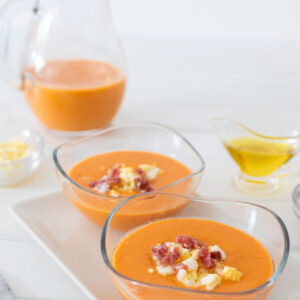 Zwei Glasschälchen cremiger orangeroter Salmorejo mit Serrano-Schinken und gehacktem Ei, dahinter ein Krug Suppe und ein Kännchen Olivenöl auf hellem Hintergrund