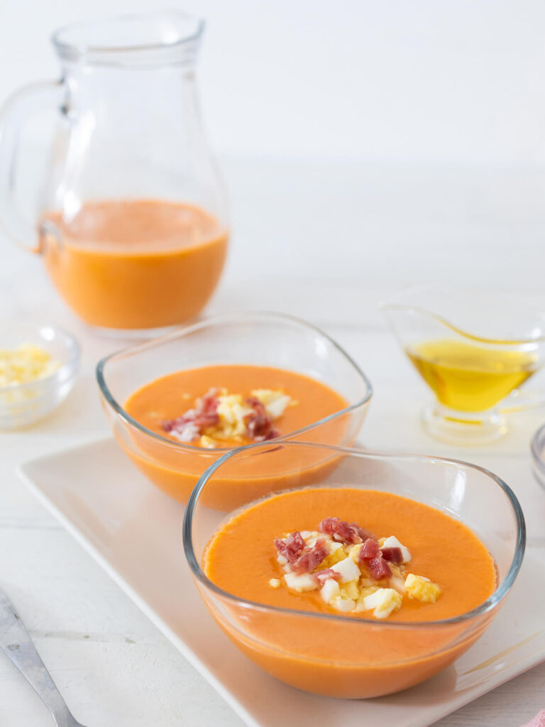 Zwei Glasschälchen cremiger orangeroter Salmorejo mit Serrano-Schinken und gehacktem Ei, dahinter ein Krug Suppe und ein Kännchen Olivenöl auf hellem Hintergrund