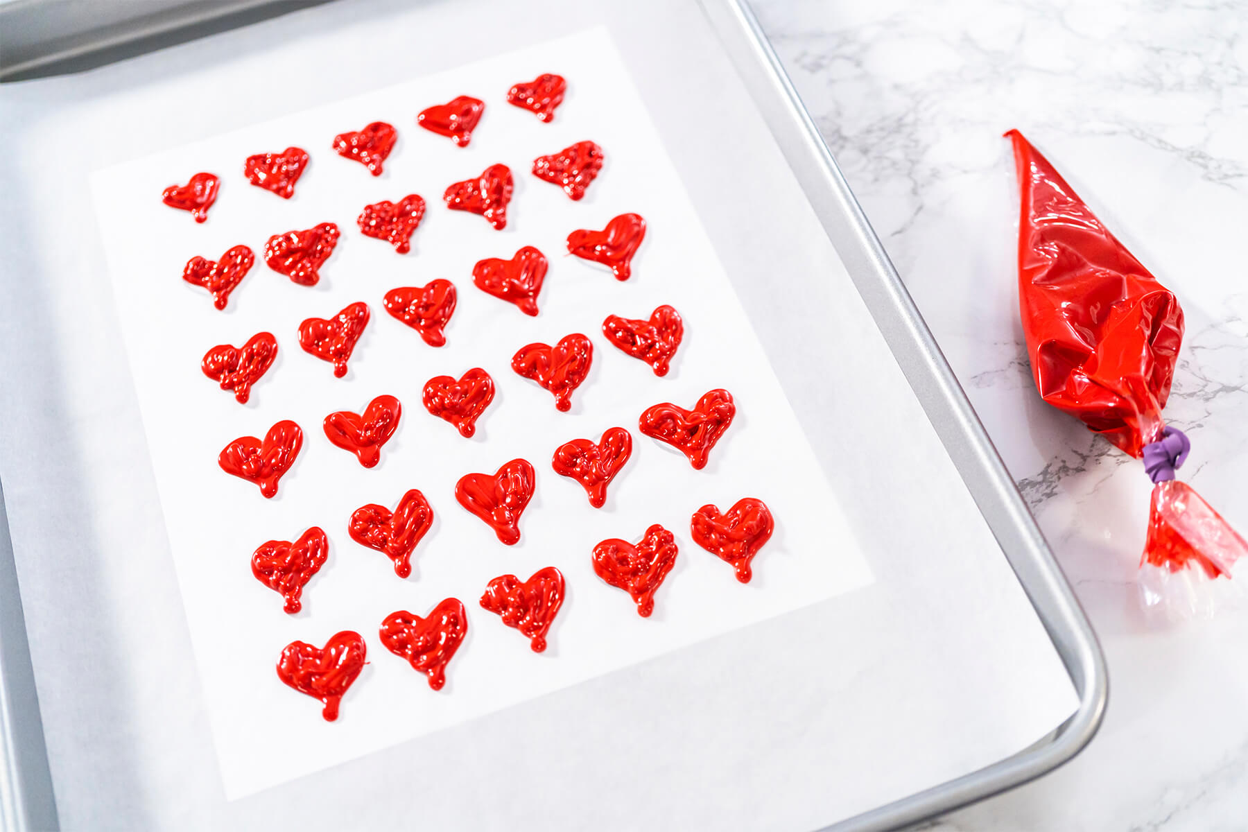 Viele fertig gespritzte rote Schoko-Herzen auf Backpapier, daneben liegt ein roter Spritzbeutel