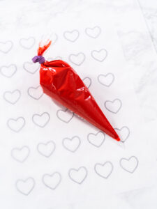 Gefüllter roter Spritzbeutel auf Backpapier über einer Vorlage mit Herz-Umrissen