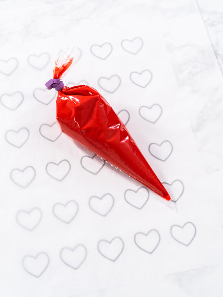 Gefüllter roter Spritzbeutel auf Backpapier über einer Vorlage mit Herz-Umrissen