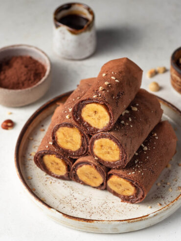 Gestapelte vegane Schoko-Crêpes-Röllchen mit Bananenfüllung, mit Kakao bestäubt und mit Haselnüssen bestreut