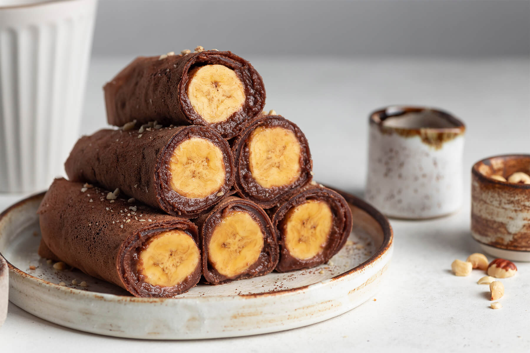 Vegane Schoko-Crêpes-Röllchen mit Bananenfüllung, aufgeschnitten und pyramidenförmig gestapelt, bestreut mit Haselnüssen