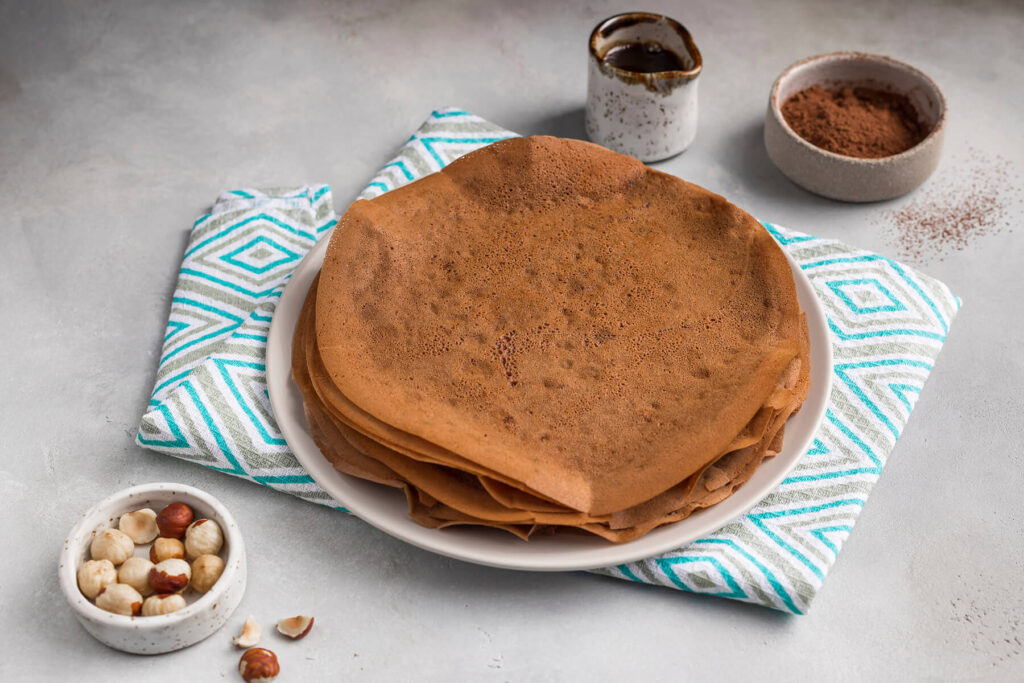 Ein Stapel dünner veganer Schoko-Crêpes auf einem Teller, daneben Kakao, Schokosauce und Haselnüsse