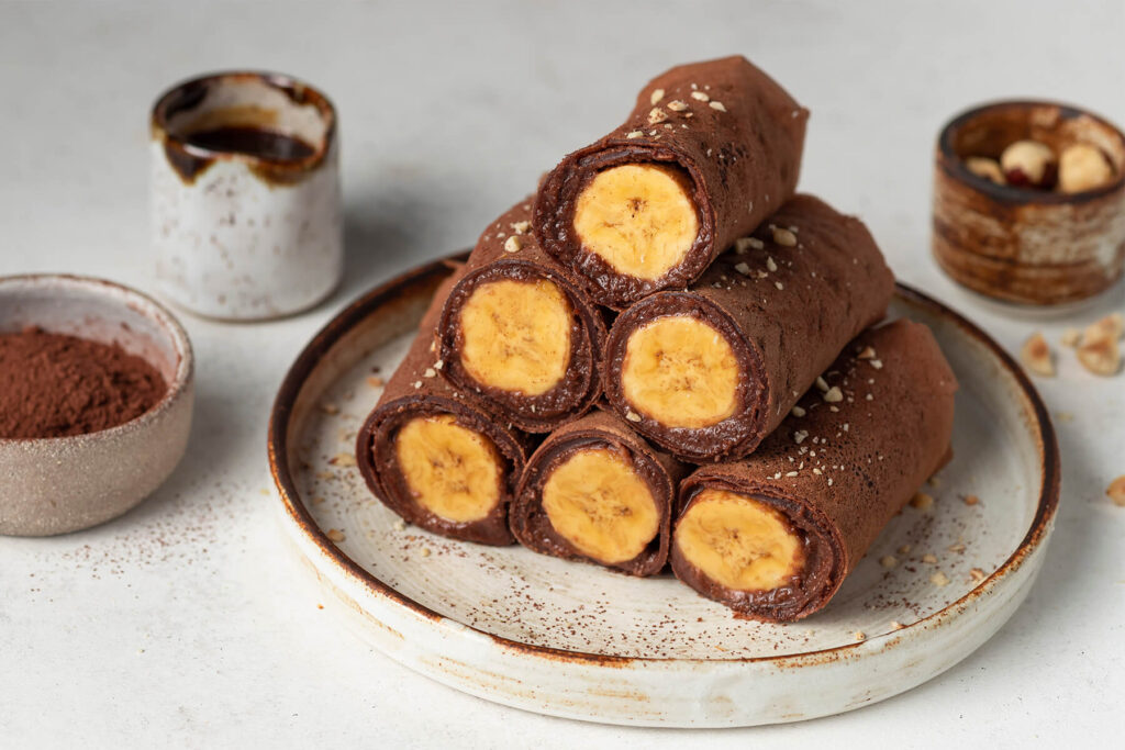 Vegane Schoko-Crêpes-Röllchen mit Bananenfüllung, angeschnitten und mit Kakao und Haselnüssen bestreut, auf einem hellen Teller