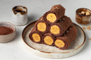 Vegane Schoko-Crêpes-Röllchen mit Bananenfüllung, angeschnitten und mit Kakao und Haselnüssen bestreut, auf einem hellen Teller