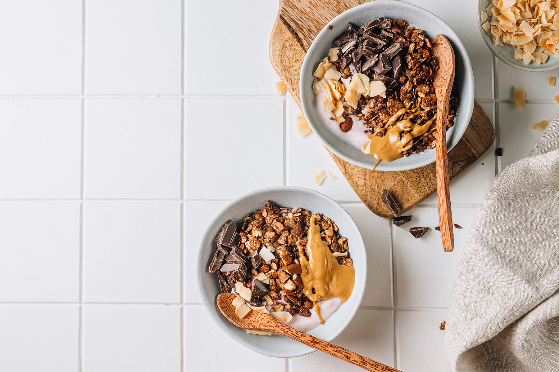 Zwei Frühstücks-Bowls mit knusprigem dunklem Schoko-Granola über rosafarbenem Beerenjoghurt, garniert mit grob gehackter dunkler Schokolade, Kokoschips und einem Löffel Erdnussmus, dazu Holzlöffel auf weißen Fliesen