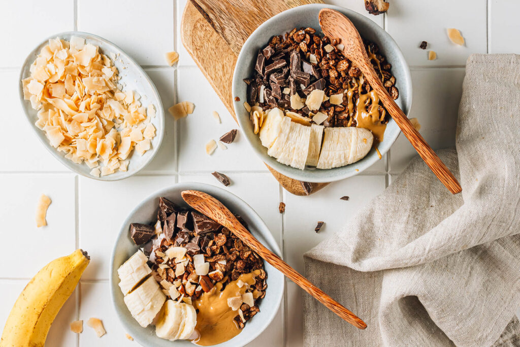Zwei fertig angerichtete Frühstücks-Bowls mit dunklem Schoko-Granola über rosafarbenem Beerenjoghurt, mit Bananenscheiben, gehackter Schokolade, Kokoschips und einem Löffel Erdnussmus, daneben eine ganze Banane und eine Schale mit Kokoschips