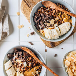 Aufsicht auf zwei Frühstücks-Bowls mit knusprigem dunklem Schoko-Granola, Bananenscheiben, grob gehackter dunkler Schokolade, Kokoschips und einem Löffel Erdnussmus über rosafarbenem Beerenjoghurt, dazu Holzlöffel auf hellen Fliesen