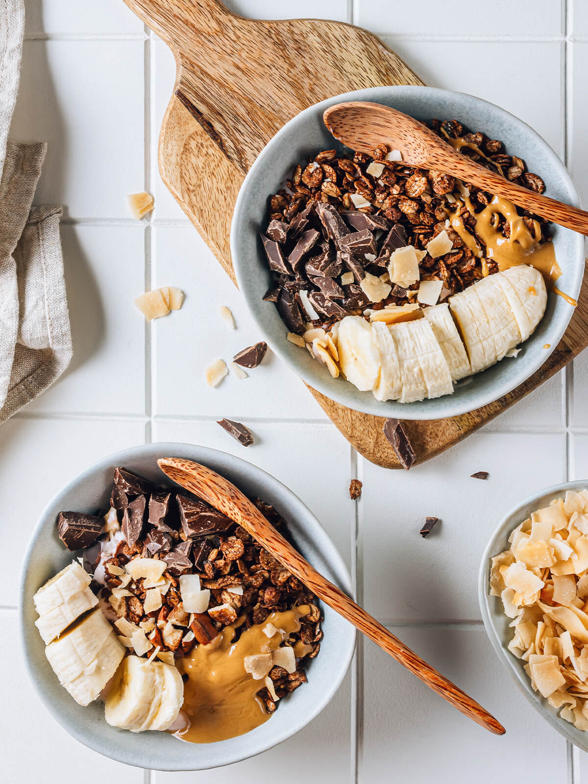 Aufsicht auf zwei Frühstücks-Bowls mit knusprigem dunklem Schoko-Granola, Bananenscheiben, grob gehackter dunkler Schokolade, Kokoschips und einem Löffel Erdnussmus über rosafarbenem Beerenjoghurt, dazu Holzlöffel auf hellen Fliesen