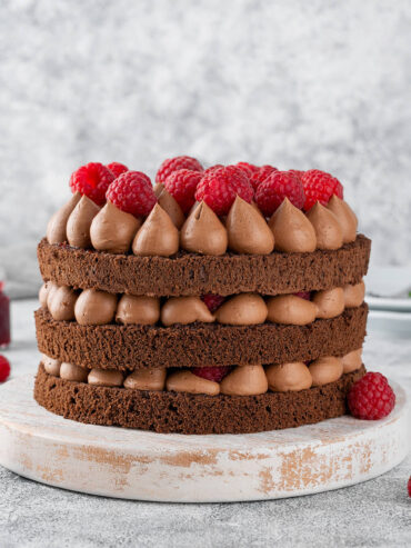 Schoko-Himbeer-Torte im Naked-Cake-Look mit drei Schokoladenbiskuits, aufgespritzten Schokocreme-Tupfen und frischen Himbeeren auf einem Holzbrett