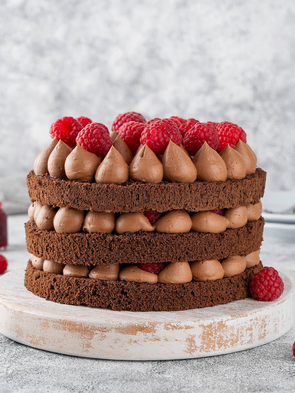 Schoko-Himbeer-Torte im Naked-Cake-Look mit drei Schokoladenbiskuits, aufgespritzten Schokocreme-Tupfen und frischen Himbeeren auf einem Holzbrett