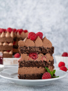 Angeschnittenes Stück Schoko-Himbeer-Torte auf einem Teller mit Schokoladenbiskuit, Schokocreme-Tupfen und frischen Himbeeren zwischen den Schichten