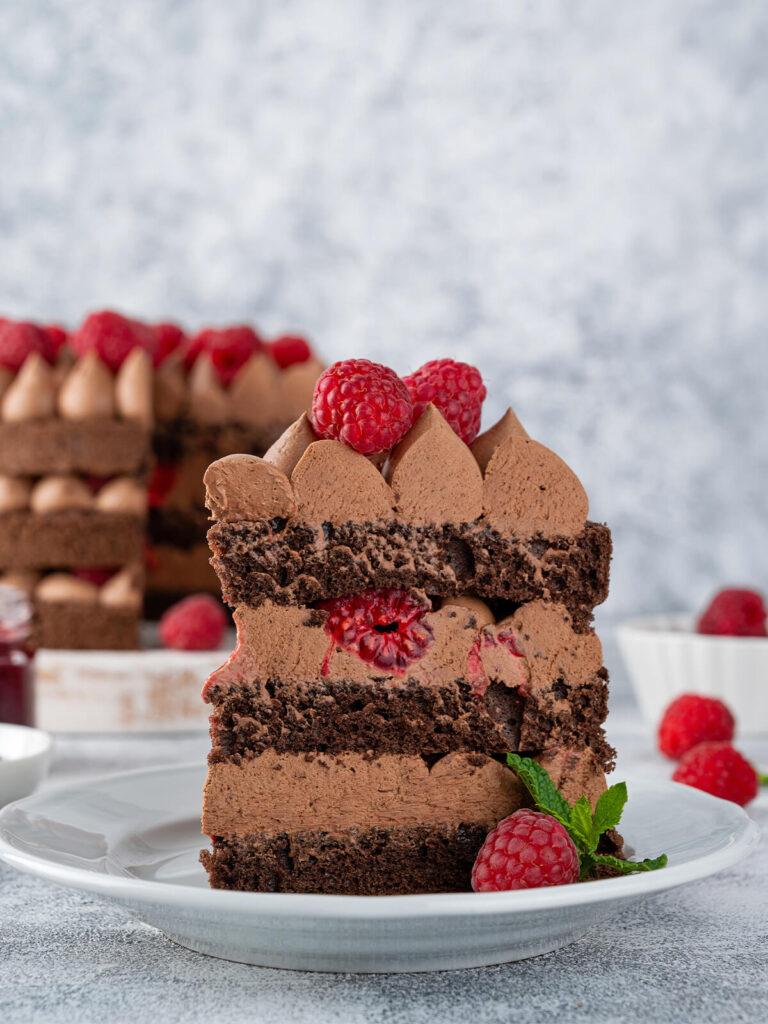 Angeschnittenes Stück Schoko-Himbeer-Torte auf einem Teller mit Schokoladenbiskuit, Schokocreme-Tupfen und frischen Himbeeren zwischen den Schichten