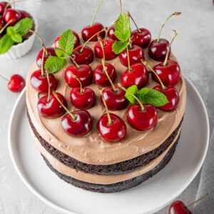 Schoko-Kirsch-Torte mit zwei dunklen Schokoladenböden, luftiger Schokosahne und frischen Kirschen mit Stiel obenauf, garniert mit Minze