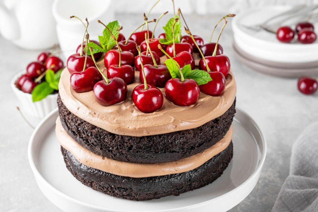 Fertige Schoko-Kirsch-Torte mit Schokosahne, dicht belegt mit ganzen frischen Kirschen mit Stiel und Minze auf einer weißen Tortenplatte