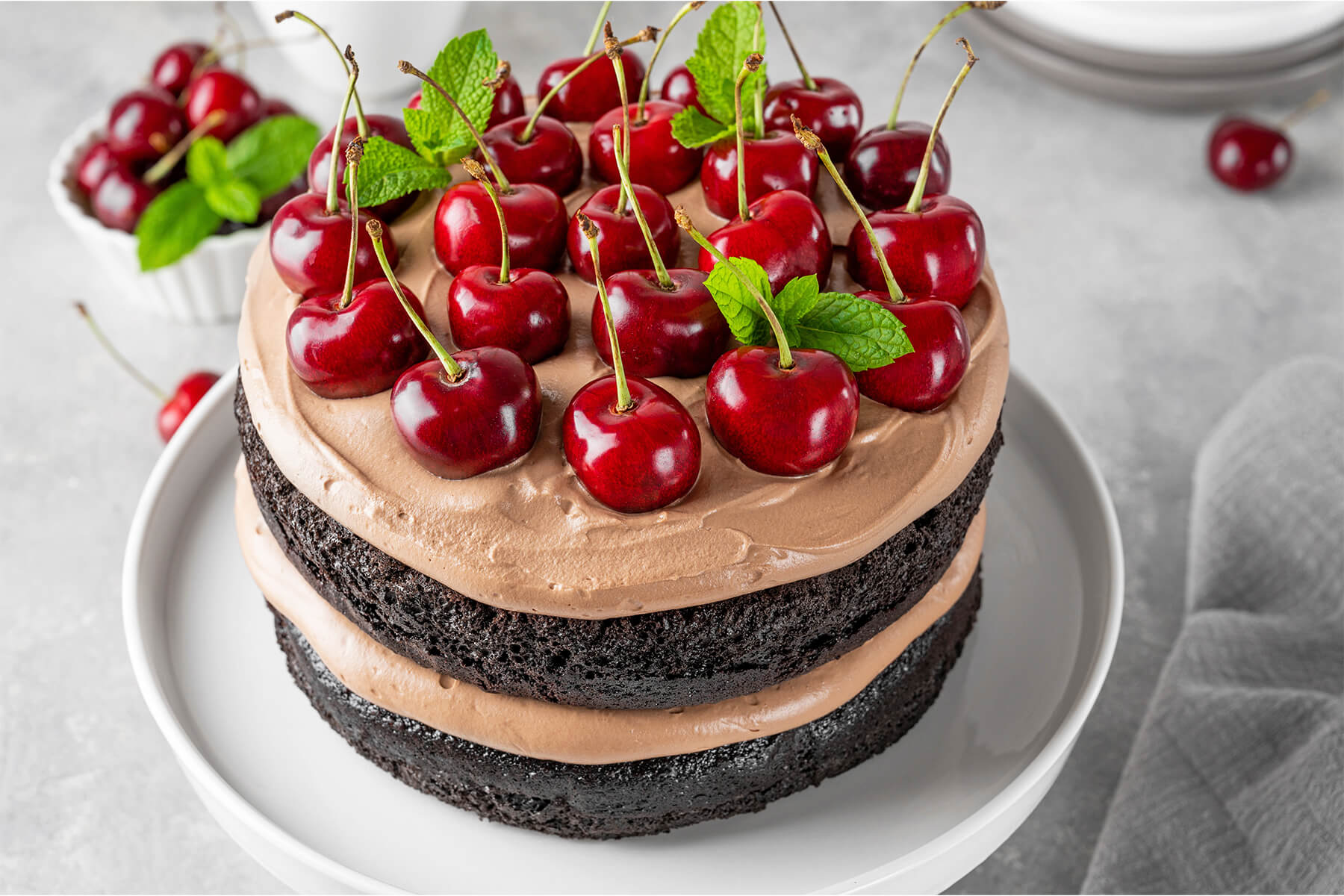 Schoko-Kirsch-Torte schräg von oben: die Schokosahne ist dicht an dicht mit ganzen frischen Kirschen mit Stiel belegt, dazwischen frische Minzblätter