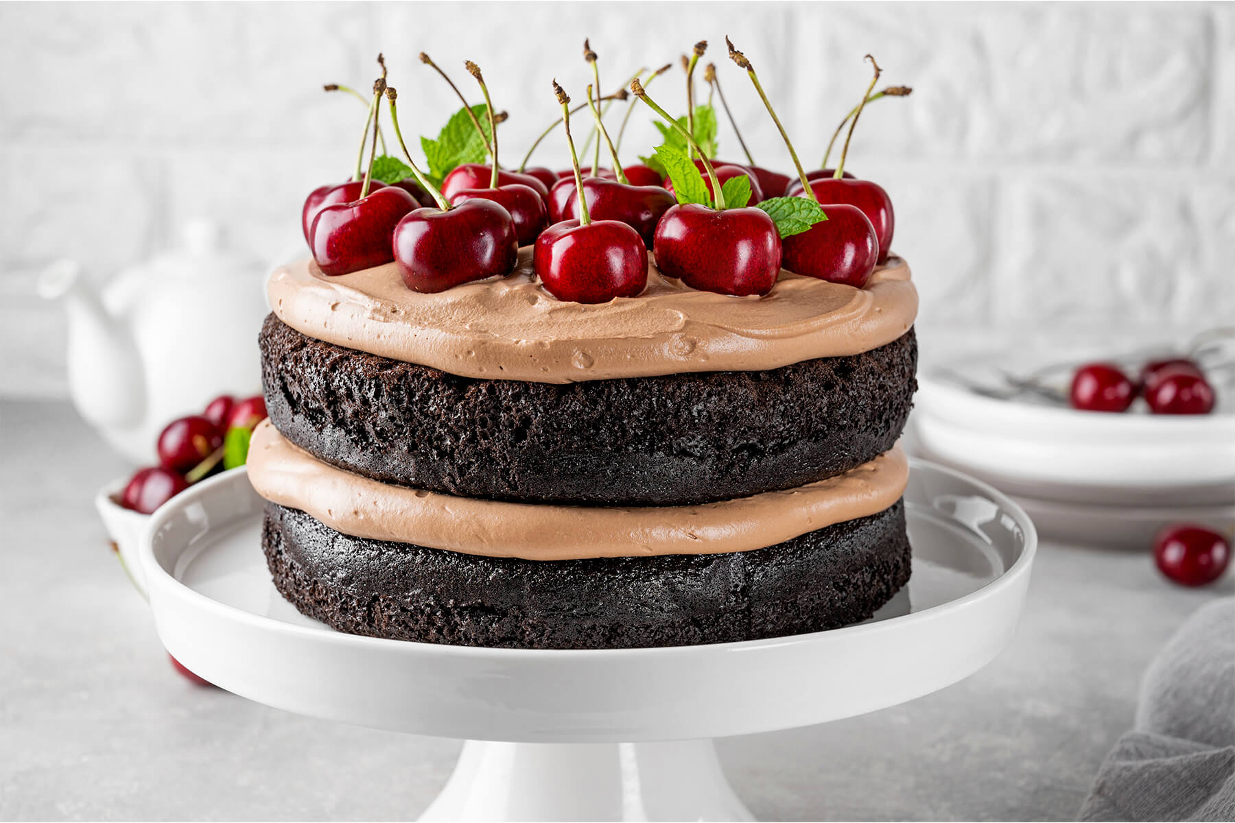 Schoko-Kirsch-Torte von der Seite auf einer weißen Tortenplatte: zwei dunkle, saftige Schokoladenböden mit einer dicken Schicht Schokosahne dazwischen und ganzen Kirschen mit Stiel obenauf