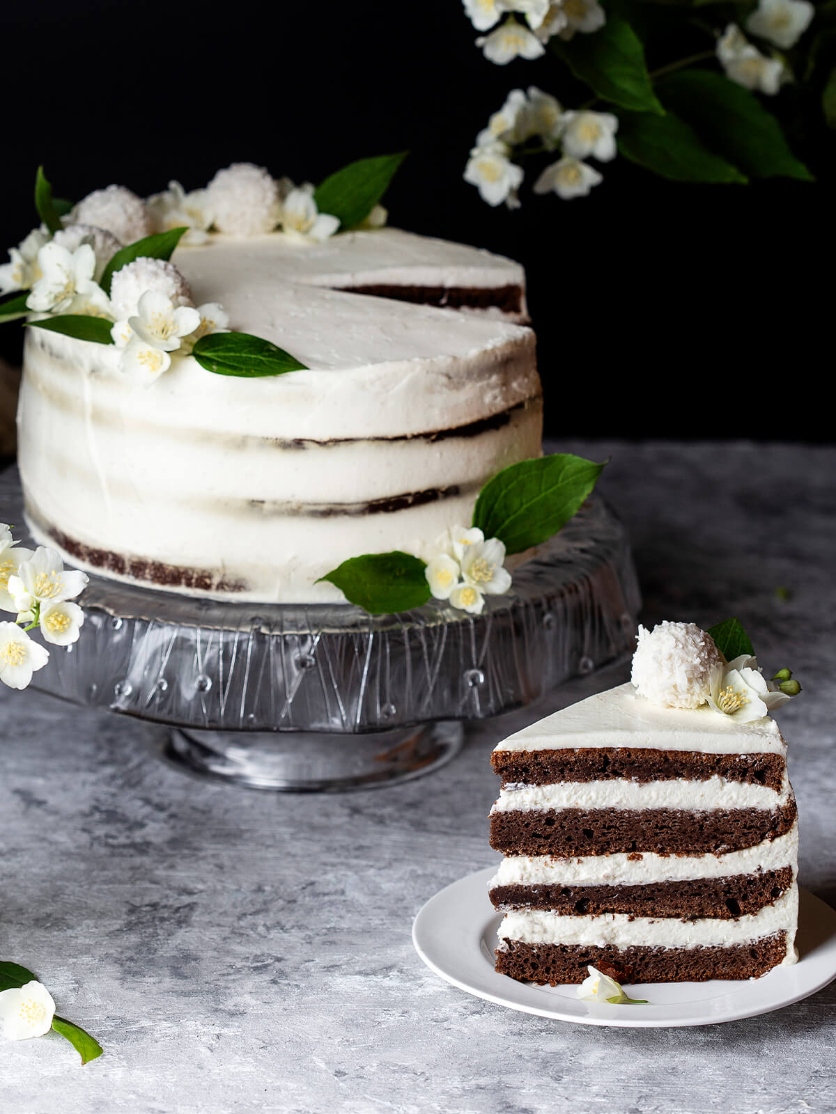 Schoko-Kokos-Torte im Naked-Cake-Stil mit weißer Creme, Kokoskugeln und Blüten, daneben ein angeschnittenes Stück mit vier dunklen Schichten