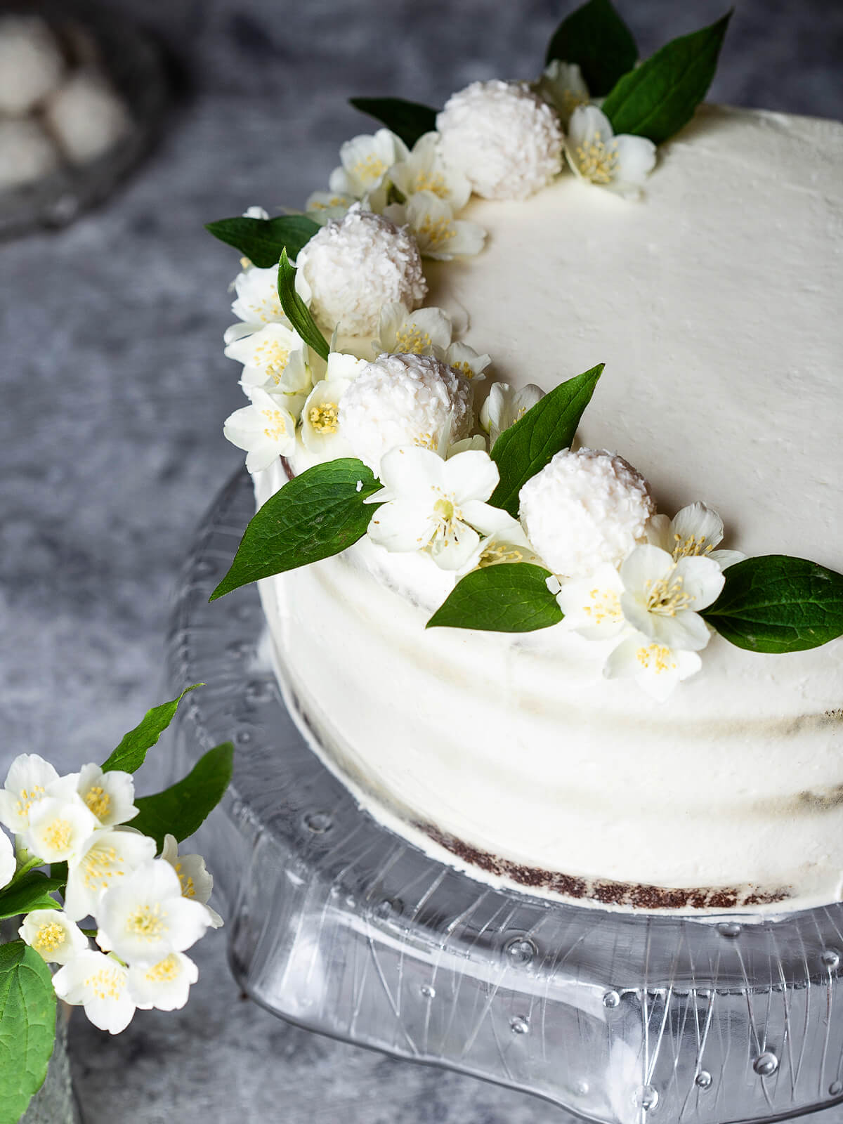 Deko der Schoko-Kokos-Torte von oben mit weißen Kokoskugeln, weißen Blüten und grünen Blättern