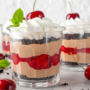 Zwei Gläser Schwarzwälder Kirsch-Trifle mit sichtbaren Schichten aus dunklen Schokoladenkeks-Bröseln, Schoko-Mascarpone-Creme und glänzender Kirschfüllung, gekrönt von einer aufgespritzten Sahnehaube mit frischer Kirsche und Minzblatt
