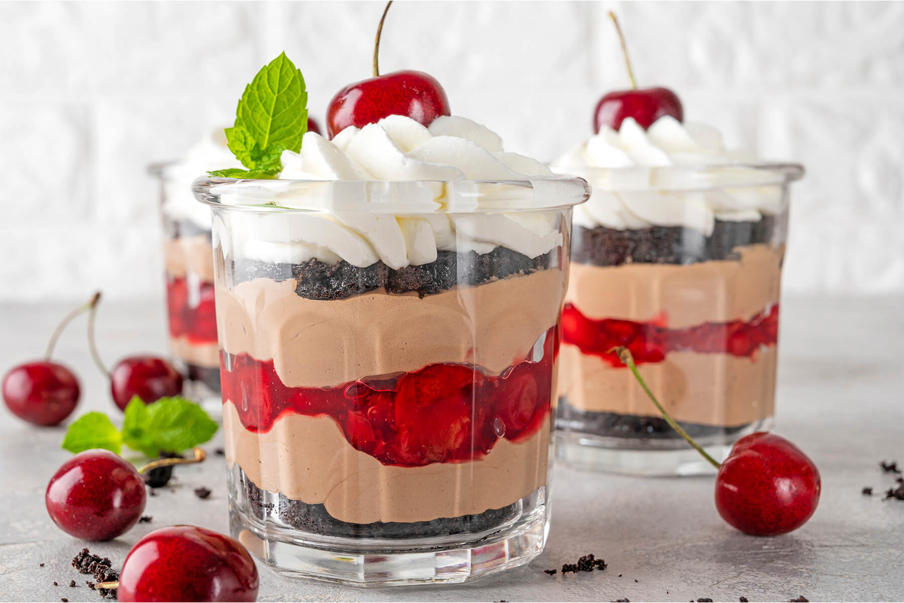 Zwei Gläser Schwarzwälder Kirsch-Trifle nebeneinander mit klar getrennten Schichten aus dunklen Schokoladenkeks-Bröseln, Schoko-Mascarpone-Creme und glänzender Kirschfüllung, gekrönt von einer Sahnehaube mit frischer Kirsche und Minze
