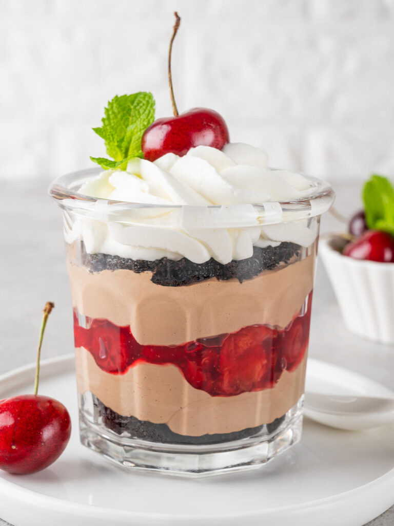 Ein Glas Schwarzwälder Kirsch-Trifle auf einem Teller mit deutlich sichtbaren Schichten aus dunklen Schokobröseln, Schoko-Mascarpone-Creme und glänzender Kirschfüllung, oben eine aufgespritzte Sahnehaube mit frischer Kirsche und Minzblatt