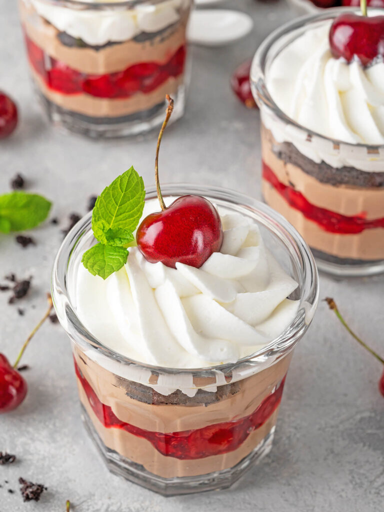 Schwarzwälder Kirsch-Trifle im Glas von oben mit luftiger, aufgespritzter Sahnehaube, einer glänzenden frischen Kirsche und einem Minzblatt, darunter die sichtbaren Schichten aus Schokocreme und Kirschen