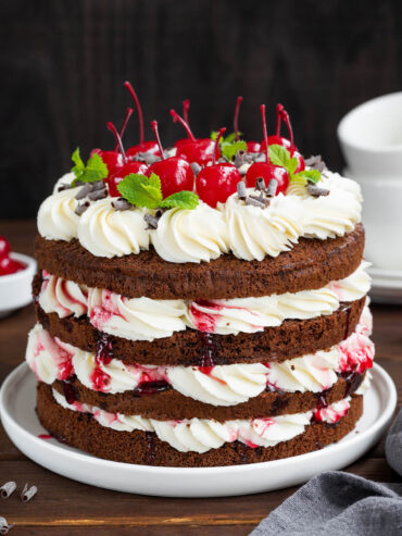 Klassische Schwarzwälder Kirschtorte im Naked-Cake-Look mit dunklen Schokoladenböden, weißer Sahne, Sauerkirschen und Cocktailkirschen auf dunklem Holztisch
