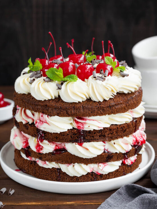 Klassische Schwarzwälder Kirschtorte im Naked-Cake-Look mit dunklen Schokoladenböden, weißer Sahne, Sauerkirschen und Cocktailkirschen auf dunklem Holztisch