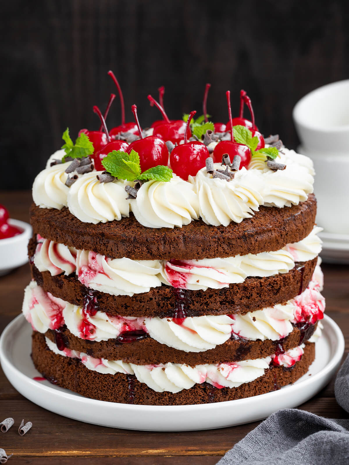 Klassische Schwarzwälder Kirschtorte im Naked-Cake-Look mit dunklen Schokoladenböden, weißer Sahne, Sauerkirschen und Cocktailkirschen auf dunklem Holztisch