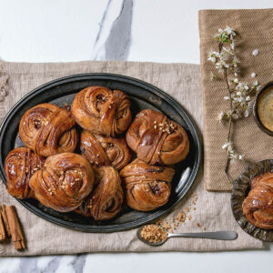 Dunkle schwedische Zimtknoten mit glänzender Oberfläche und grobem Rohrzucker, angerichtet mit Kaffee und Zimtstangen