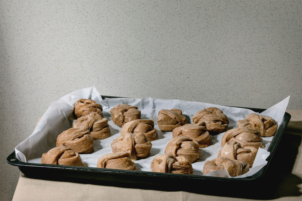 Geschlungene Knoten aus dunklem Zimtteig mit Abstand auf einem mit Backpapier belegten Backblech