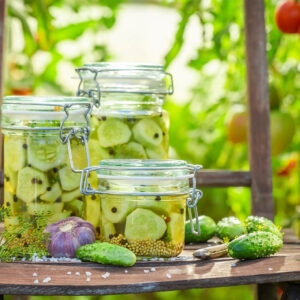 Mehrere Gläser mit süß-sauer eingelegten Gurkenstücken, Senfkörnern, Piment und Dill auf einem Tisch im Garten