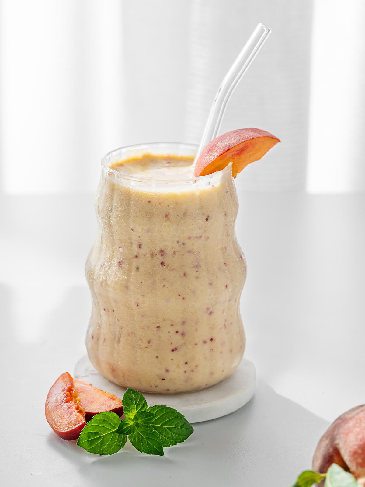 Cremiger Pfirsich-Bananen-Smoothie im Glas mit Glasstrohhalm, Pfirsichspalte und Minze