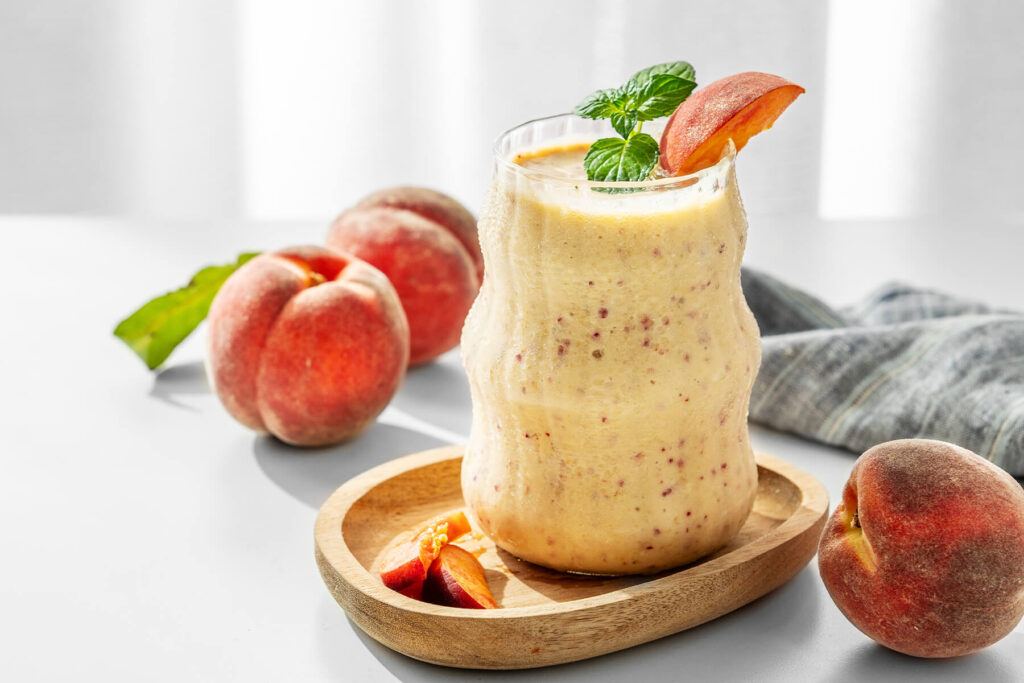 Cremiger Pfirsich-Bananen-Smoothie im Glas mit Minze und Pfirsichspalte auf einem Holzteller, daneben frische Pfirsiche