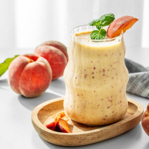 Cremiger Pfirsich-Bananen-Smoothie im Glas mit Minze und Pfirsichspalte auf einem Holzteller, daneben frische Pfirsiche