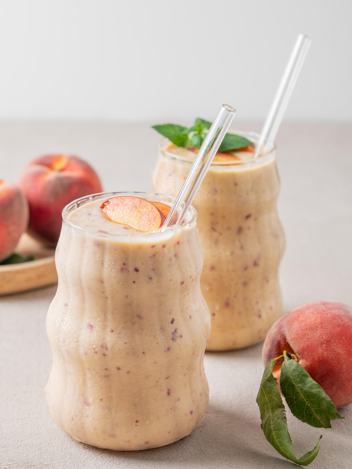 Zwei Gläser cremiger Pfirsich-Bananen-Smoothie mit Glasstrohhalmen, daneben frische Pfirsiche