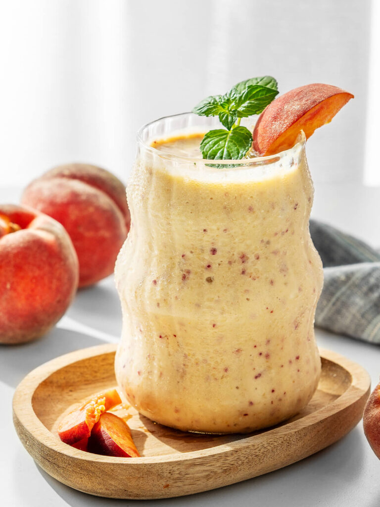 Einzelnes Glas Pfirsich-Bananen-Smoothie auf einem Holzteller, garniert mit Minze und Pfirsich