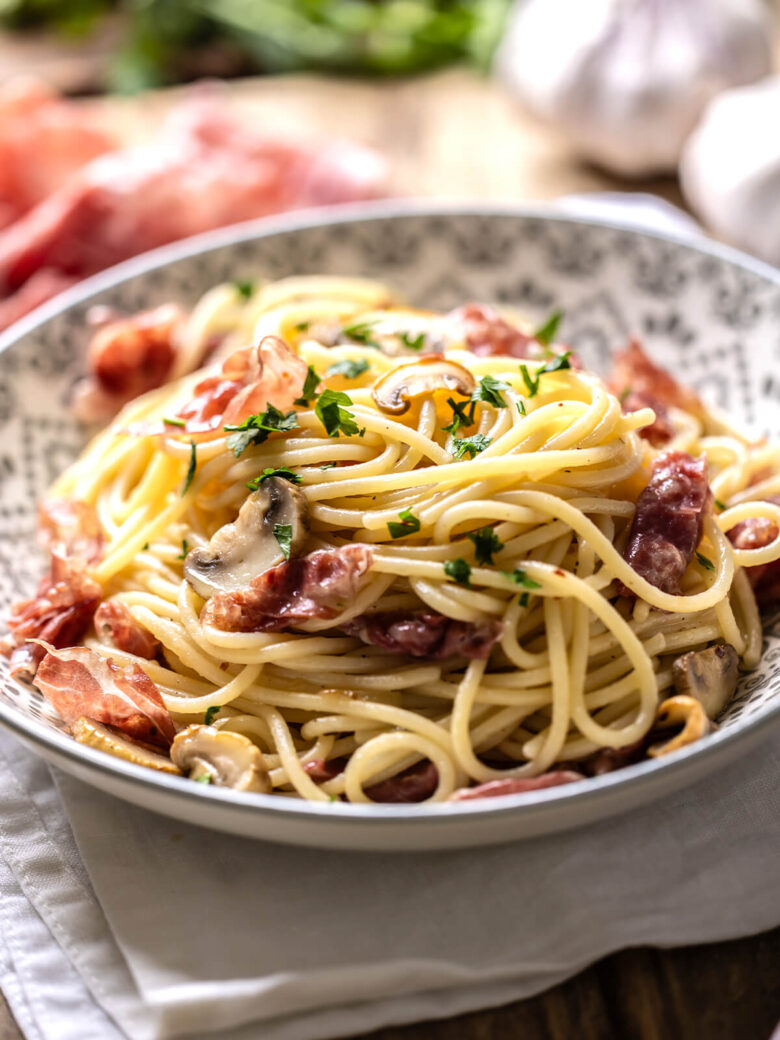 Spaghetti mit Prosciutto, Champignons und Petersilie in einer gemusterten Schale, im Hintergrund Rohschinken und Knoblauch